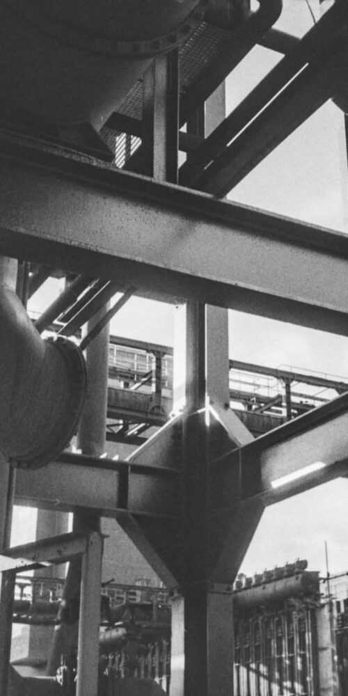 Black and white view of an industrial facility with steel beams, large pipes, and structural framework seen from below.