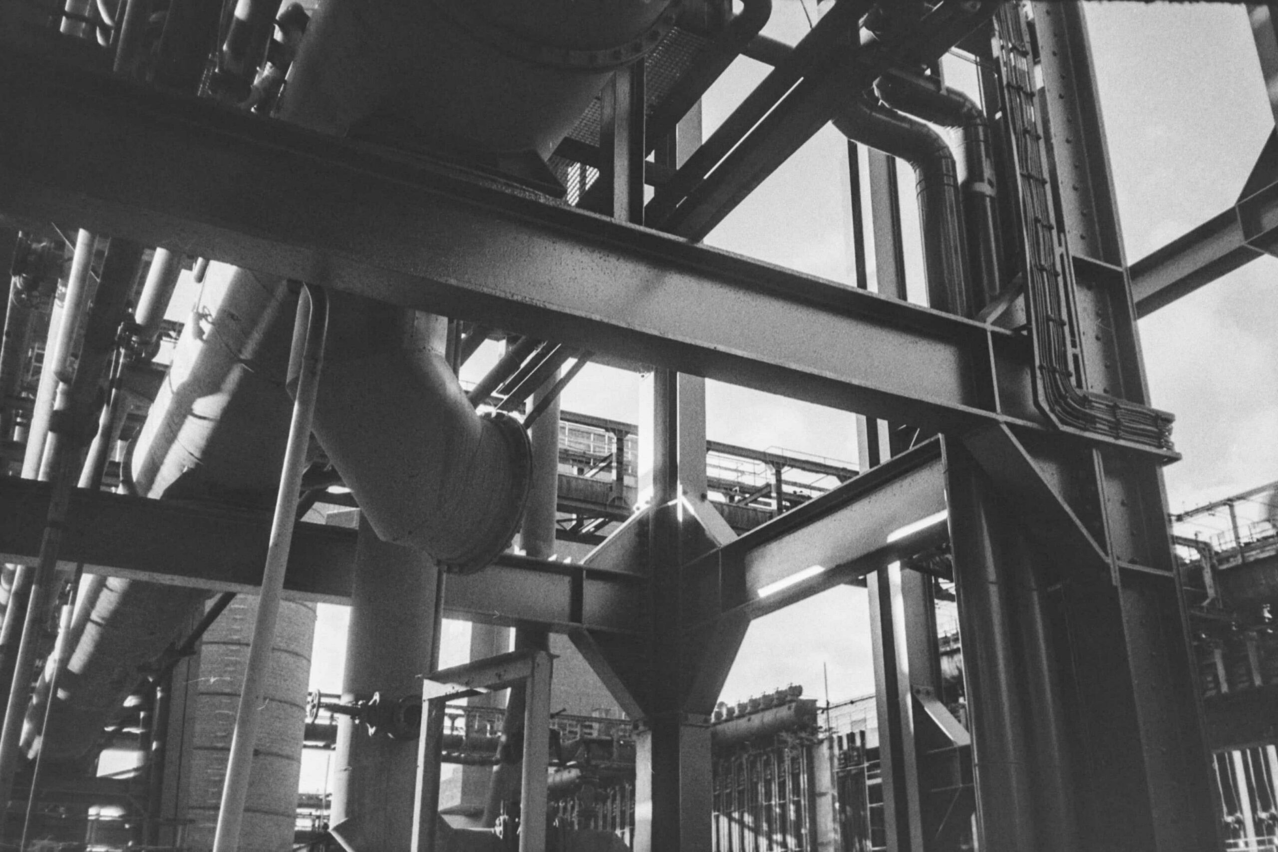 Black and white view of an industrial facility with steel beams, large pipes, and structural framework seen from below.