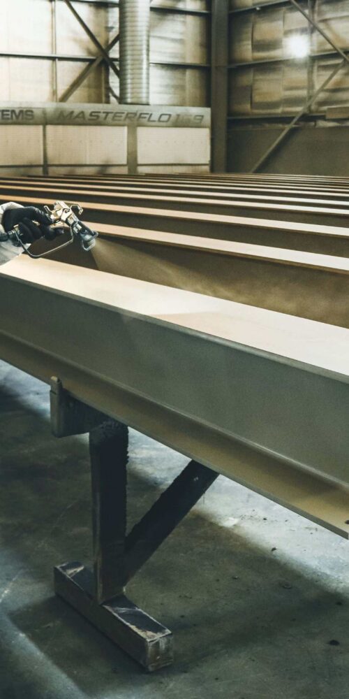Steel fabrication shop worker applying protective coating to structural steel beams for industrial manufacturing project